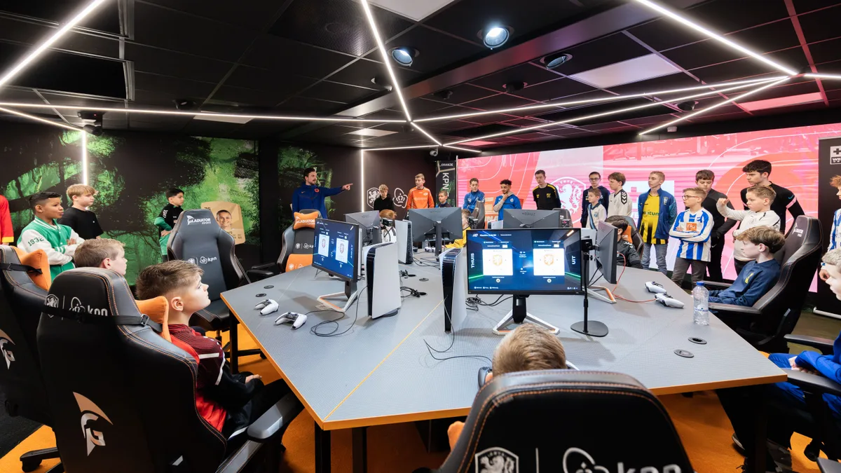Breed overzicht van de esports room met jongeren in voetbalshirts rondom de gametafel met PS5-stations