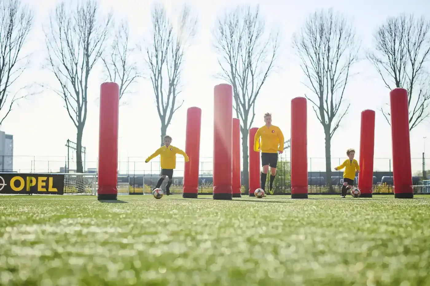 Drie jeugdspelers in gele shirts dribbelen met de bal tussen opblaasbare rode zuilen op een voetbalveld