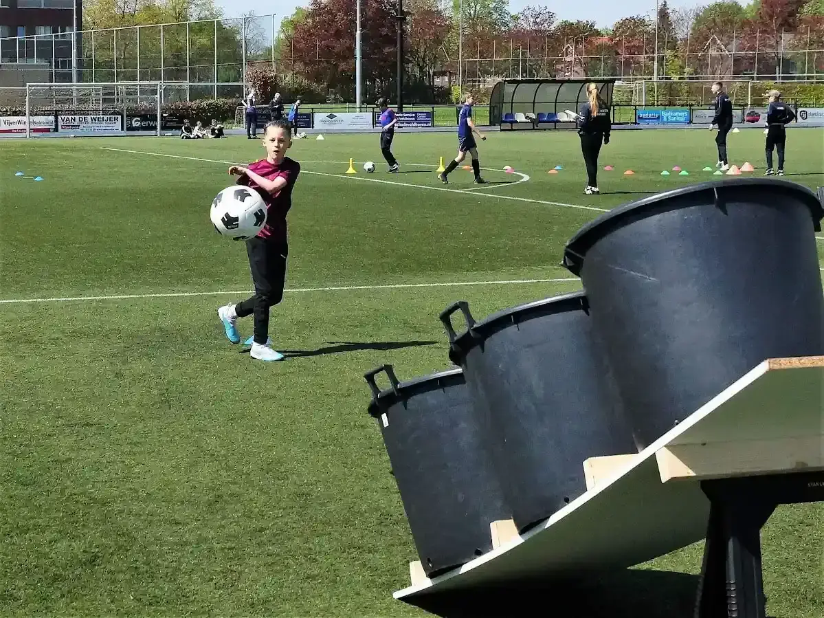 Kind gooit voetbal richting emmers op een schuin platform terwijl andere spelers oefenen op het veld