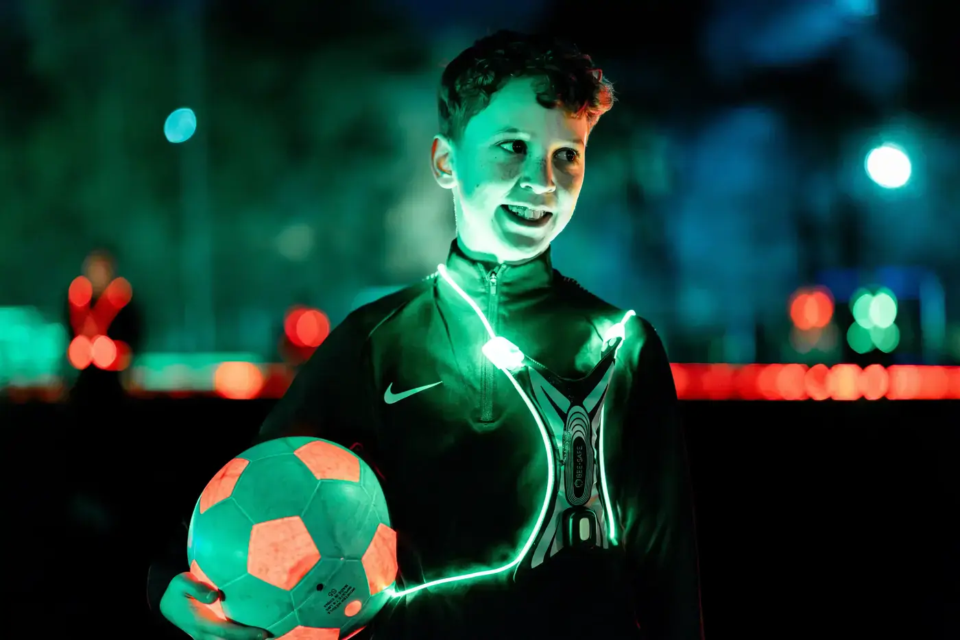 Lachende jongen houdt een oplichtende voetbal vast en draagt een LED-hesje tijdens een avondwedstrijd
