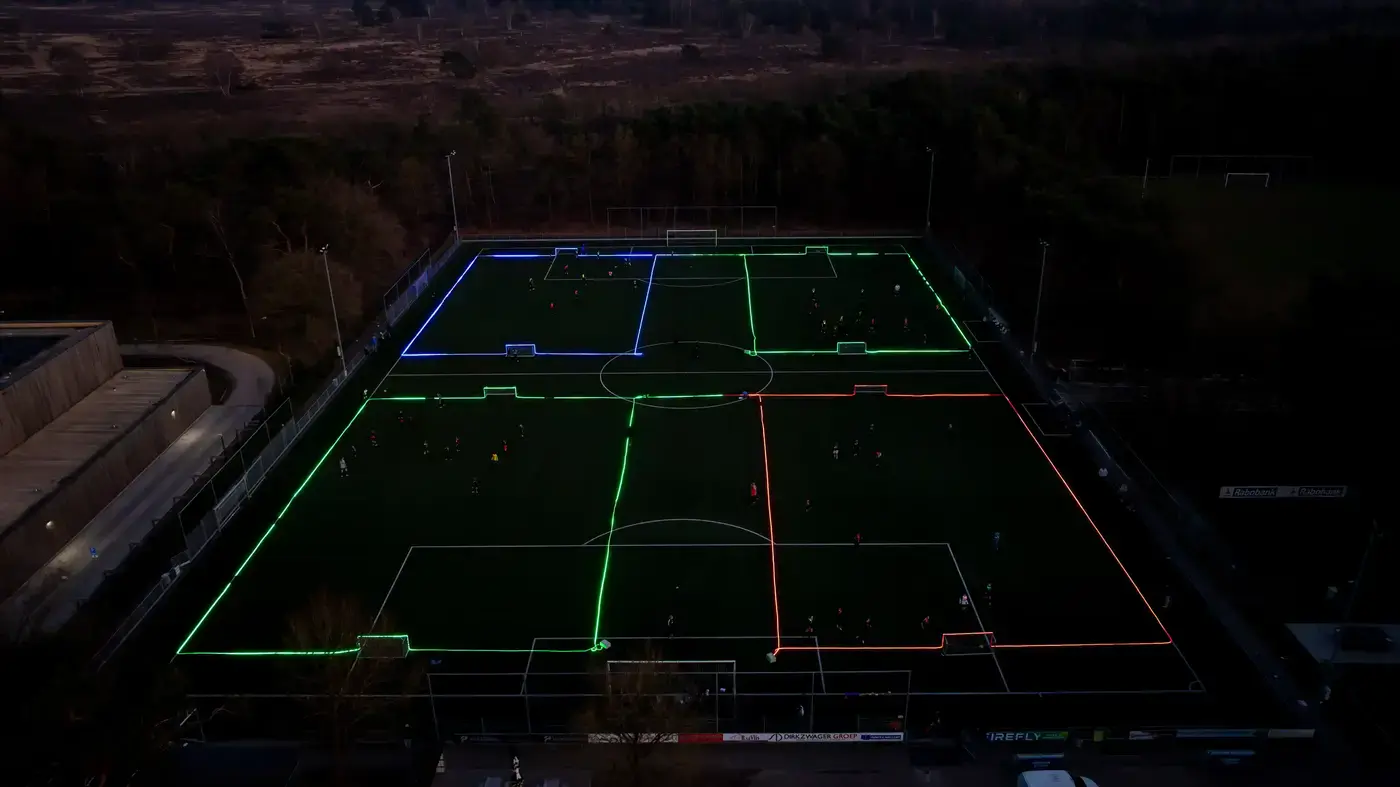 Dronefoto van een groot sportveld verdeeld in LED-verlichte speelvelden met spelers in actie bij schemering