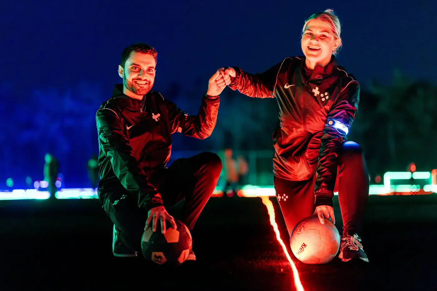 Twee lachende begeleiders knielen met voetballen op een LED-verlicht veld met neonlijnen op de achtergrond